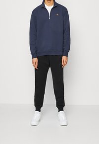 Granatowa bluza z półzamkiem i małym logo na piersi, w zestawieniu z czarnymi spodniami dresowymi. Buty są białe z teksturowanym wzorem.