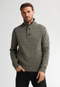 Bărbat îmbrăcat cu un pulover tricotat ribbed de culoare verde măsliniu, cu guler înalt și două nasturi, stând cu o mână în buzunar pe un fundal simplu.