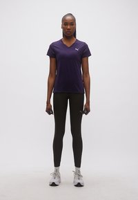 Γυναίκα όρθια κρατώντας μικρούς αλτήρες, φορώντας μωβ πουκάμισο, μαύρο κολάν, λευκές κάλτσες και αθλητικά παπούτσια σε απλό φόντο.