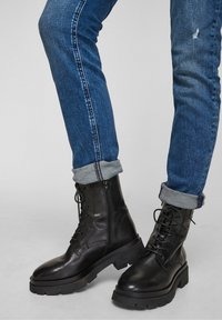 Botas de combate de cuero negro con una suela de goma resistente, punta redondeada y cordones; combinadas con jeans de mezclilla azul remangados en los puños.