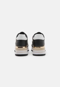 Sneakers bianco e nero con un tacco accentato in oro, suola a strati e design elegante. La parte superiore testurizzata include materiali lisci e perforati.