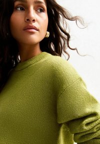 Pull en tricot vert avec un col rond et des poignets côtelés ; texture douce avec de légères variations de teinte. Les cheveux encadrent doucement le visage.
