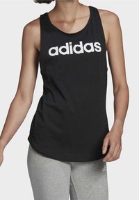Svart ärmlös linne med en stor vit "adidas"-logotyp centrerad på bröstet. Materialet verkar mjukt med en slät textur.