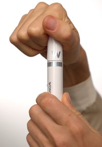 Weißer zylindrischer Applikator mit silbernen Akzenten. Hand drückt den oberen Teil, mit "W"-Logo. Glatte Oberfläche, ergonomisches Design.