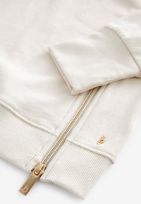 Beiger Sweatshirt mit Rippbündchen und Saum, versehen mit einem goldenen Reißverschluss und einem kleinen gestickten Logo-Detail am Bund. Aus weichem Baumwollmaterial.