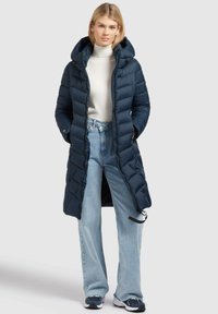 Marinblå puffjacka med huva, kviltat mönster, framdragkedja, två sidfickor, bärs över en vit polotröja och blå jeans.