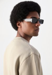 Occhiali da sole neri rettangolari con una montatura nera lucida e tempie in metallo argento a contrasto. Finitura liscia e testurizzata. Modello che indossa un felpa beige.