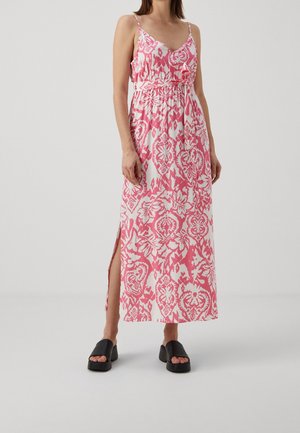 Kvinde iført en ærmeløs, lang kjole med pink og hvidt mønster og sideslids samt sorte plateau-sandaler, stående mod en ensfarvet baggrund.