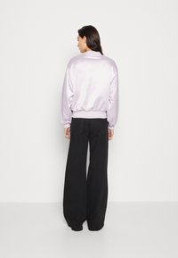 Giacca bomber in satin lilla con orlo e polsini a costine, abbinata a jeans neri a gamba larga. La giacca presenta una texture liscia e lucida.