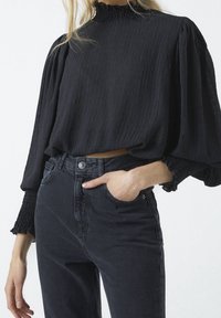 Schwarze, strukturierte, verkürzte Bluse mit langen, gepufften Ärmeln und rüschiertem Ausschnitt. Kombiniert mit dunklen, hoch sitzenden Jeans. Hand-in-Tasche-Detail.
