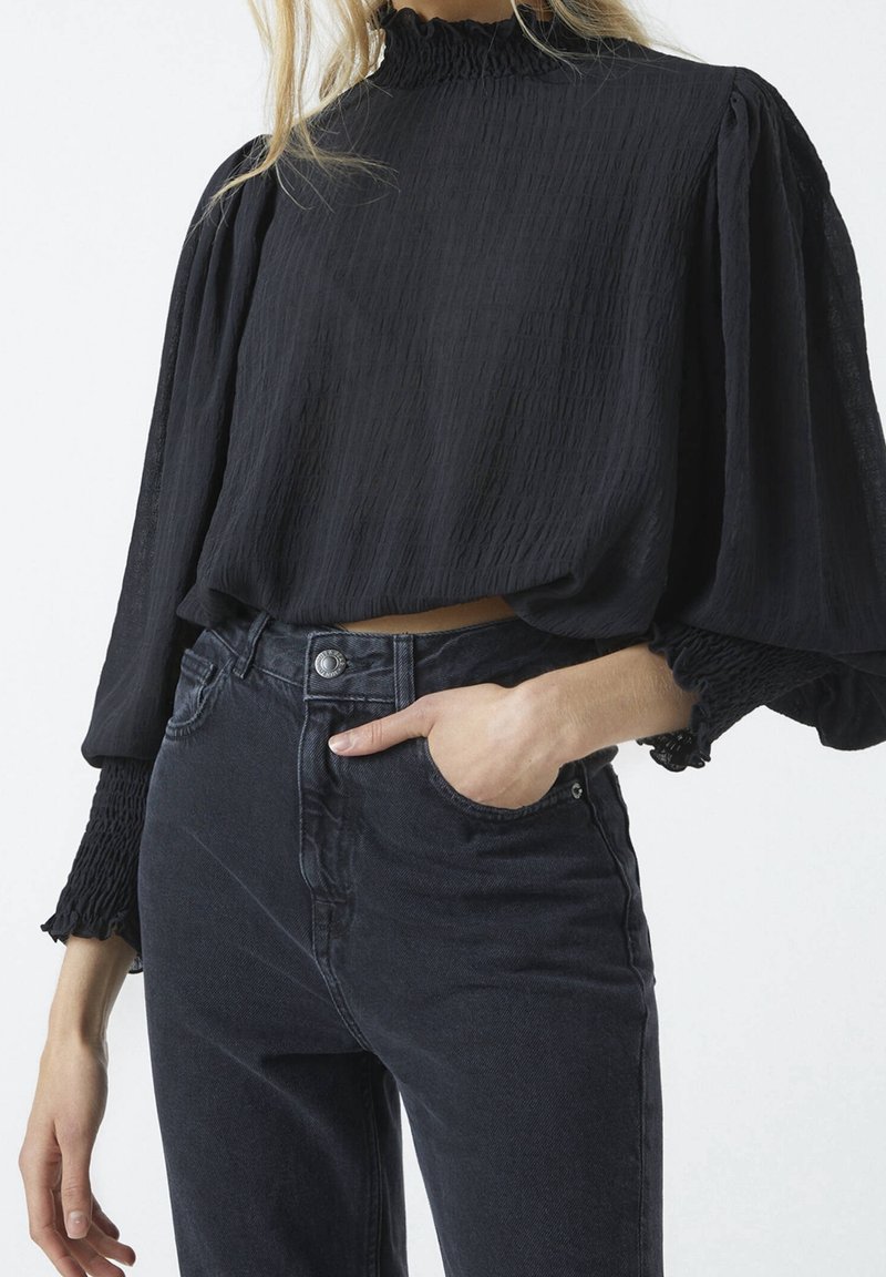 Schwarze, strukturierte, verkürzte Bluse mit langen, gepufften Ärmeln und rüschiertem Ausschnitt. Kombiniert mit dunklen, hoch sitzenden Jeans. Hand-in-Tasche-Detail.