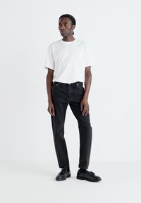 Jeans negre din denim cu croi drept, asortați cu un tricou alb simplu, cu guler pe gât. Modelul poartă pantofi negri, pozând pe un fundal alb.