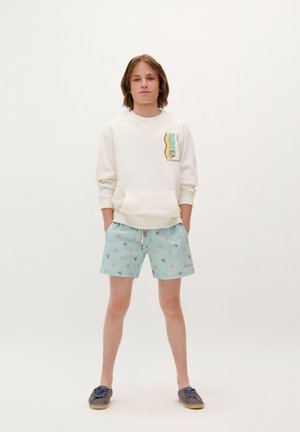 Felpa color crema chiaro con stampa grafica, abbinata a pantaloni da bagno verde menta con motivi floreali. Le scarpe espadrillas marroni completano il look.