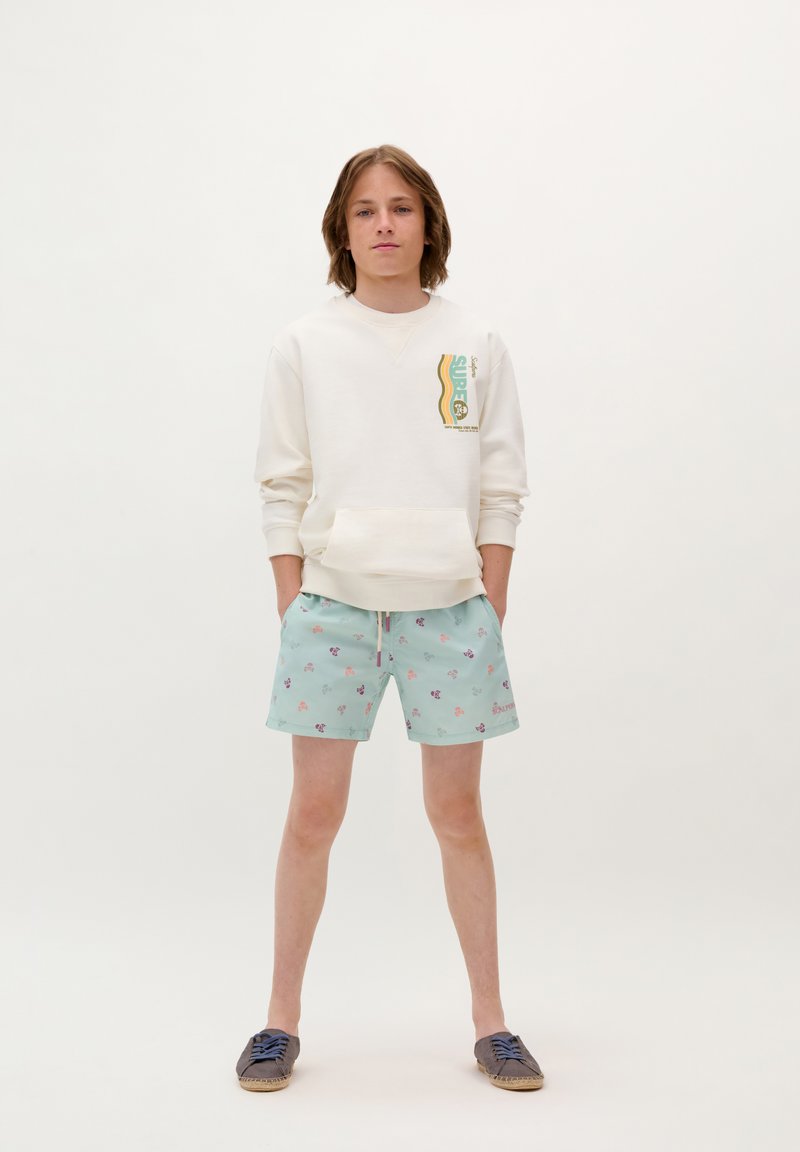 Lichte crèmekleurige sweatshirt met grafische print, gecombineerd met mintgroene zwemshorts met bloemenpatronen. Bruine espadrille schoenen maken de look compleet.