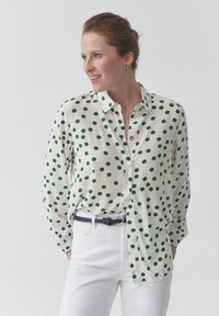 Camicia bianca con pois verdi, maniche lunghe, leggero lucido, abbinata a pantaloni bianchi e una cintura nera. Nessun motivo prominente sui pantaloni.