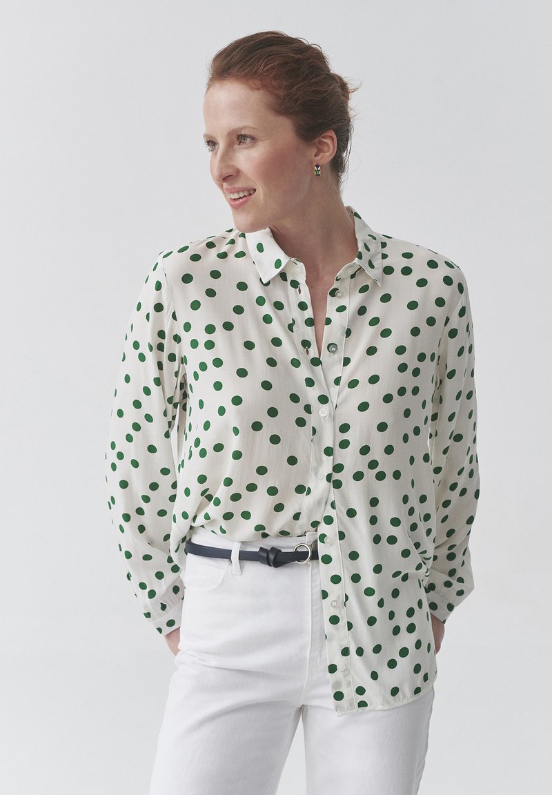 Camicia bianca con pois verdi, maniche lunghe, leggero lucido, abbinata a pantaloni bianchi e una cintura nera. Nessun motivo prominente sui pantaloni.