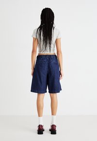 Shorts de mezclilla en azul oscuro, con un diseño casual, cinturilla elástica y dobladillos enrollados, combinados con una camiseta gris ajustada y zapatos negros.