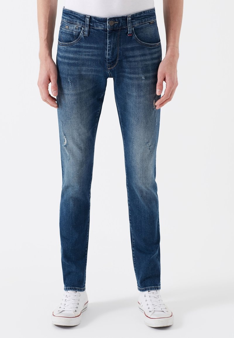 Homme portant un jean bleu slim avec une légère usure et des baskets blanches sur un fond blanc uni.