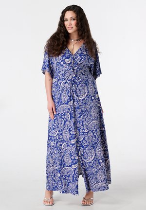 Vrouw met lang krullend haar, gekleed in een blauwe en witte maxi-jurk met bloemenpatroon en zilveren sandalen, staand tegen een effen achtergrond.