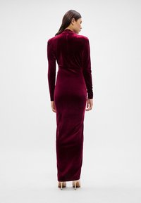 Μακρύ βυσσινί βελούδινο φόρεμα με μακριά μανίκια και εφαρμοστή σιλουέτα, με ψηλό λαιμόκομμα και σχέδιο που φτάνει μέχρι το πάτωμα.