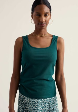 Frau trägt ein ärmelloses, dunkelgrünes Oberteil und einen blauen Rock mit weißem Blumenmuster, goldene Creolen und eine dünne goldene Halskette.