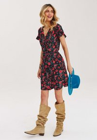 Květinové šaty s krátkým rukávem v černé barvě, zdobené červenými a zelenými květinovými vzory. V kombinaci s béžovými volnými botami a modrou čepicí.