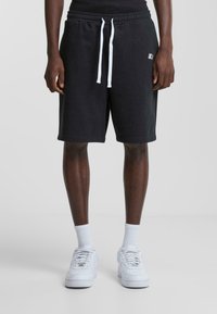 Pantalones cortos de forro polar negros con cintura elástica y cordones blancos. Presenta un pequeño logotipo en el muslo izquierdo. Se llevan con zapatillas blancas.