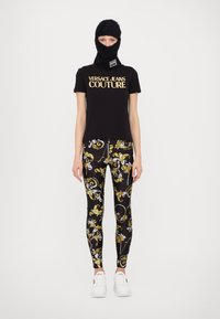 Juodas trumpomis rankovėmis marškinėliai su auksiniu "VERSACE JEANS COUTURE" logotipu, derinami su raštuotomis leggingomis, turinčiomis juodus, auksinius ir baltus raštus, ir baltais sportbačiais.