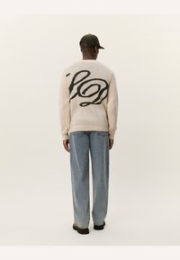 Cremig gestrickter Pullover mit einem großen schwarzen, geschwungenen Logo auf dem Rücken, kombiniert mit lockeren, hellen Jeans und einer dunkelgrünen Mütze.