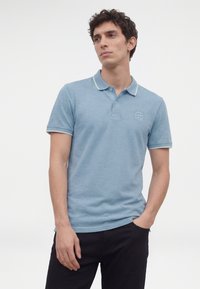 Tânăr cu păr creț și închis la culoare, purtând un tricou polo bleu deschis și pantaloni negri, stând cu o mână în buzunar, pe fundal alb.