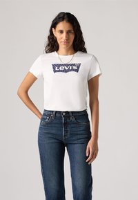 Balta medvilninė marškinėliai su tamsiai mėlynu Levi's logotipo raštu, dekoruotu taškeliais, derinami su tamsiai mėlynomis džinsinėmis aukštos talpos kelnėmis. Trumpomis rankovėmis, klasikinio kirpimo.