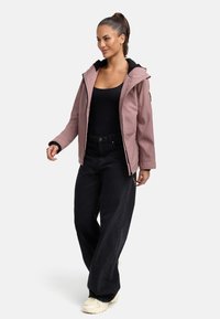 Pink softshell jacket with a zip front, black hood lining, and side pockets. Paired with black wide-leg pants and beige footwear.