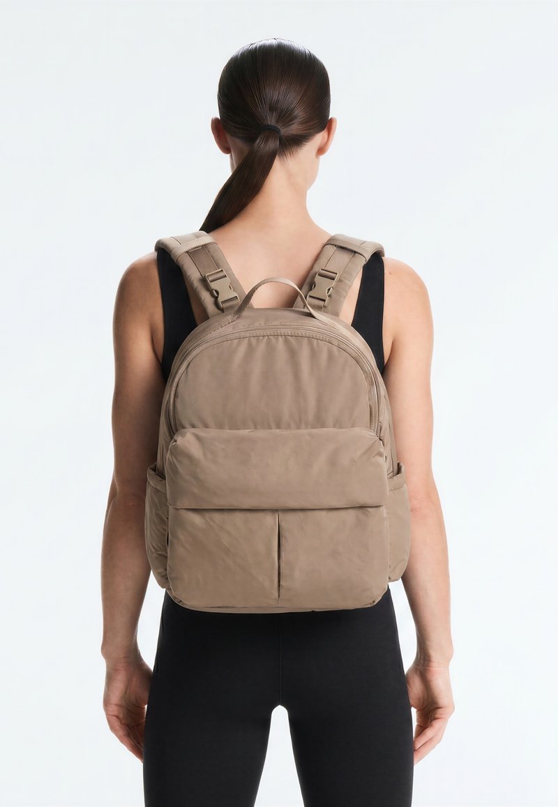 Frau mit dunklem Haar im Pferdeschwanz, die schwarze Kleidung und einen beigen Rucksack trägt, von hinten vor einem einfarbigen hellen Hintergrund.