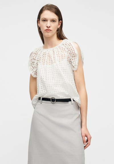 Femme portant un chemisier blanc à manches bouffantes en œillet, rentrée dans une jupe gris clair avec une ceinture noire, debout devant un fond blanc uni.