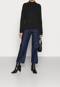 Zwarte ribgebreide trui met een hoge hals, gecombineerd met wijde donkerdenim jeans en zwarte enkellaarsjes met hak, en een zwarte tas vasthoudend.