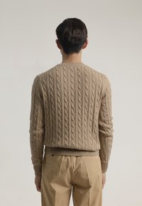 Suéter beige de punto trenzado con cuello redondo y dobladillo acanalado, que presenta un patrón texturizado. Combinado con pantalones de color claro.