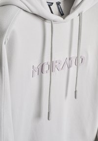 Sudadera gris claro hecha de tela suave; cuenta con un logo frontal en bordado gris más claro, cordones y paneles laterales acanalados.