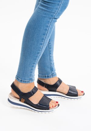 Person wearing blue jeans and black open-toe sandals with white soles, red painted toenails, standing on a white surface.