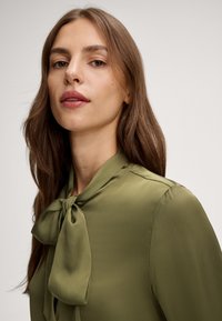 Blusa verde oliva con colletto a fiocco, texture fluida e maniche lunghe. Il tessuto appare liscio e leggero.