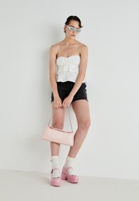 Weißes, gerafftes, schulterfreies Oberteil, schwarze Shorts, rosa Plateauschuhe, weiße Kniestrümpfe, hält eine rosa Handtasche mit Kettenriemen.