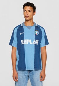 Mėlynos ir šviesiai mėlynos spalvų blokų trumpomis rankovėmis futbolo marškiniai su apykakle, kairėje krūtinės pusėje esantis "REPLAY" logotipas ir herbas.
