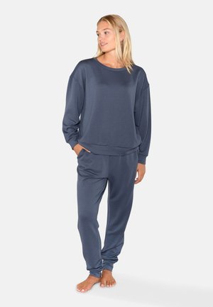 Vrouw staat op blote voeten, draagt een loszittend donkergrijs sweatshirt met lange mouwen en bijpassende joggingbroek, handen in de zakken, kijkt naar rechts.