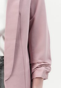 Blazer rose clair texturé avec manches retroussées et encolure dégagée, fabriqué en tissu lisse, dévoilant partiellement une chemise blanche en dessous.