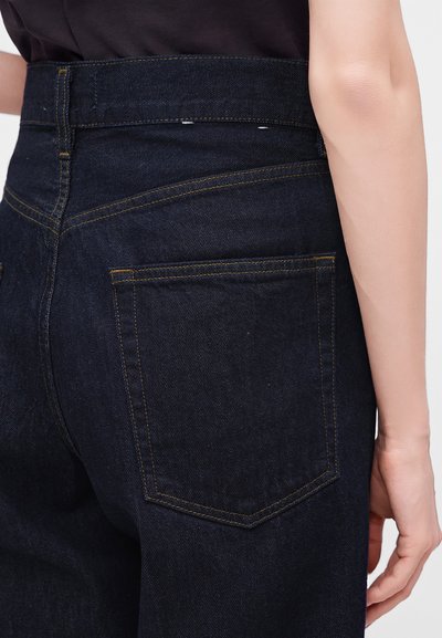 Gros plan d'une personne portant un jean en denim bleu foncé montrant la poche arrière et le côté de la hanche et du bras sur un fond clair.