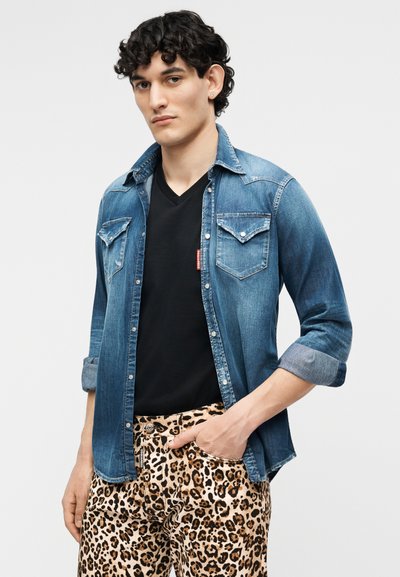 Νέος άνδρας με γυρισμένο το τζιν πουκάμισο πάνω από μαύρο μπλουζάκι V λαιμόκοψη και παντελόνι με λεοπάρ σχέδιο, στέκεται με το ένα χέρι στην τσέπη.