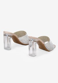 Mules de tacón transparente con una amplia tira embellecida en plata y base beige. Presentan un tacón bloque transparente y un forro texturizado en dorado.