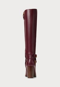 Alto stivale in pelle bordeaux con tacco in legno impilato e fascia decorativa con fibbia dorata sul retro.