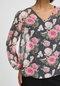 Gennemsigtig bluse med V-hals, med et blomstermønster af pink roser og hvide kallaliljer på en mørkegrå baggrund. Lange, puffede ærmer.