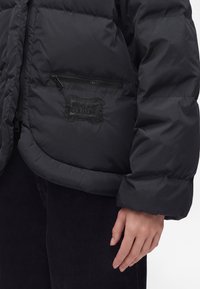 Veste matelassée noire avec poche zippée et écusson en relief "Versace Jeans Couture" à l'avant, portée sur un pantalon noir, main détendue sur le côté.