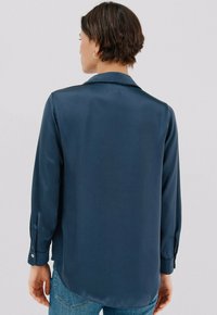 Blusa di seta blu navy con collo a camicia, maniche lunghe e polsini abbottonati. Texture liscia e orlo posteriore leggermente allungato.
